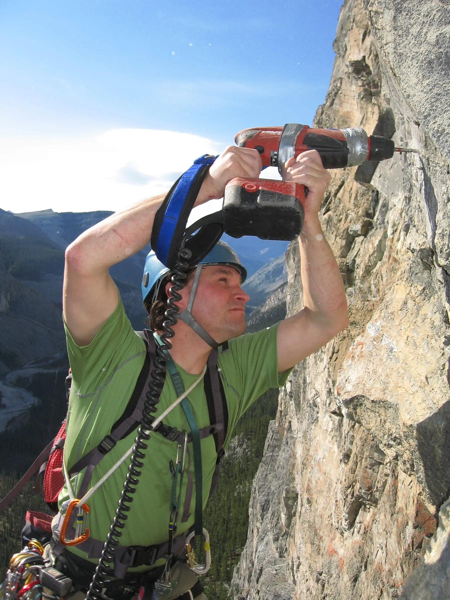 A route developer in a blue helmet drills overhead into a limestone wall, with a river valley far below