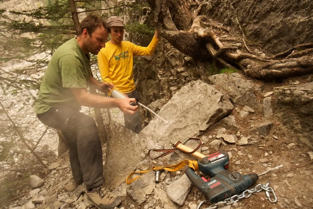 Two climbers at the foot of a cliff, one injecting glue into a hole drilled in a boulder while the other watches, with a drill and chain on the ground