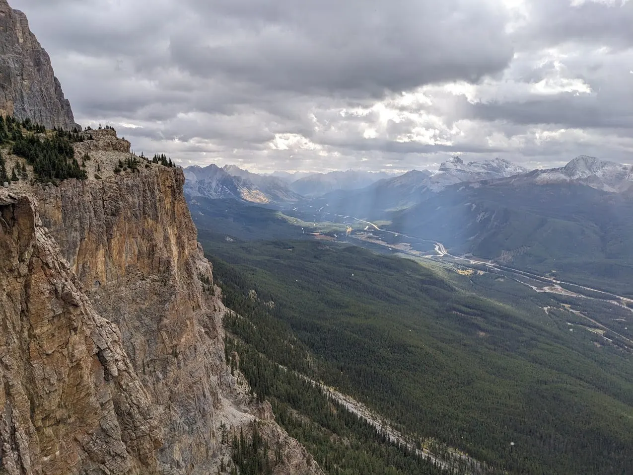 A tall limestone cliff above a forested valley, with sun breaking through clouds over the mountains