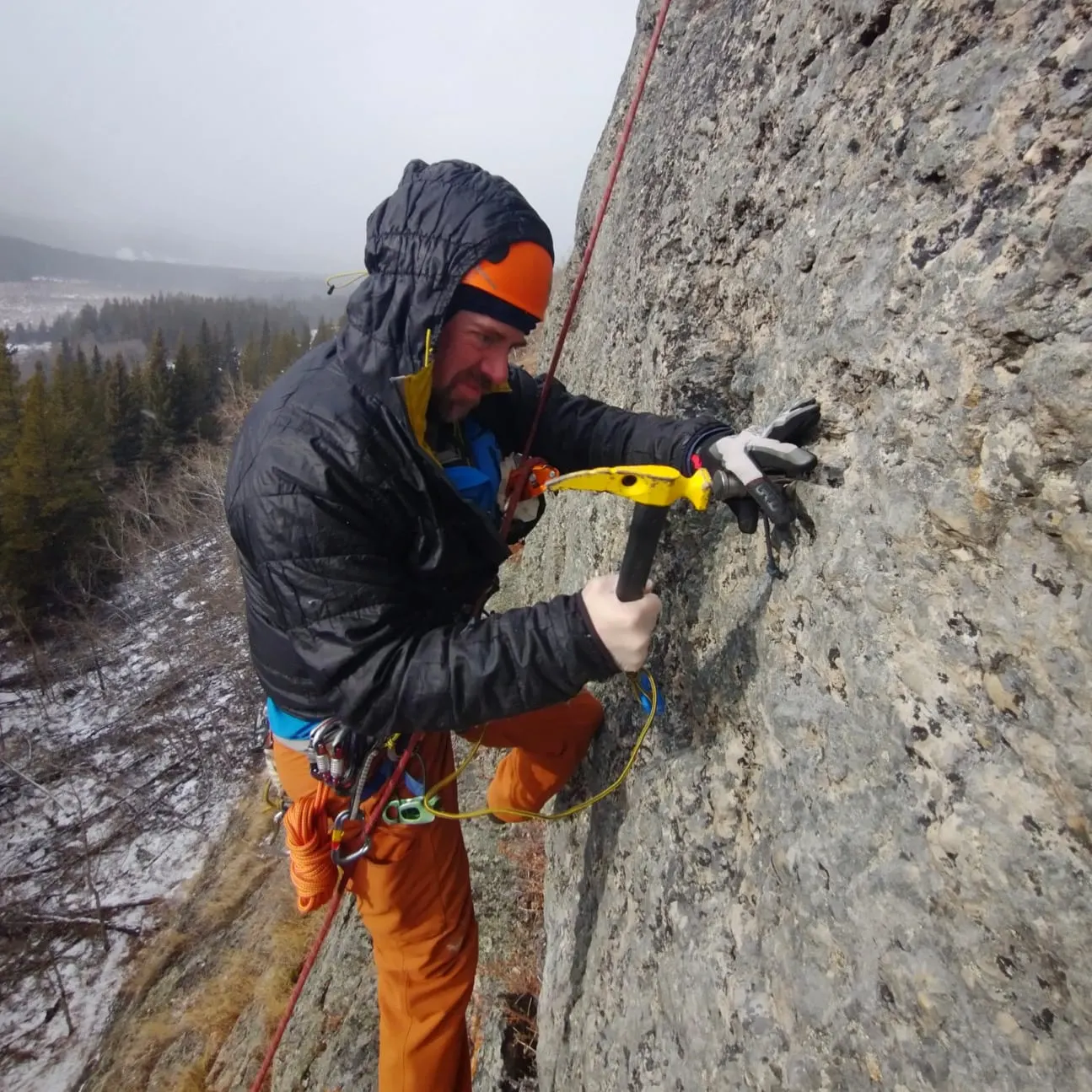 A climber in an orange hat and hooded jacket hammers a glue-in bolt into a conglomerate wall
