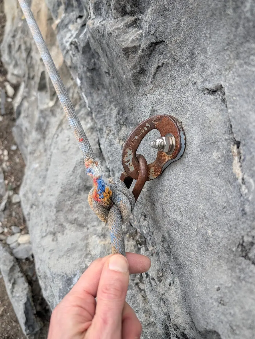 A rusted bolt hanger on grey limestone, with an old knotted rope threaded through it