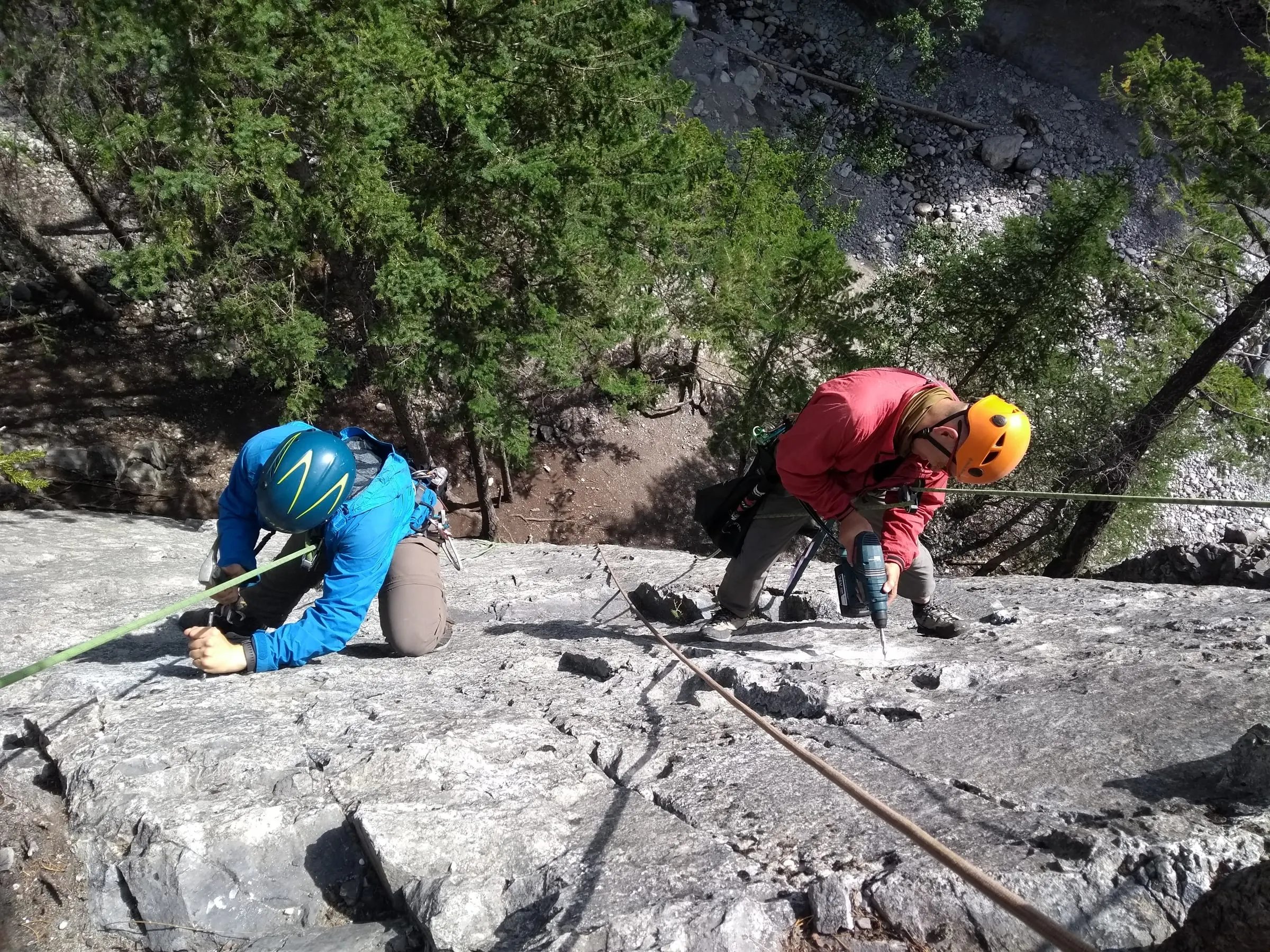 Two volunteers kneel on a limestone slab replacing bolts, one drilling while the other works on a bolt