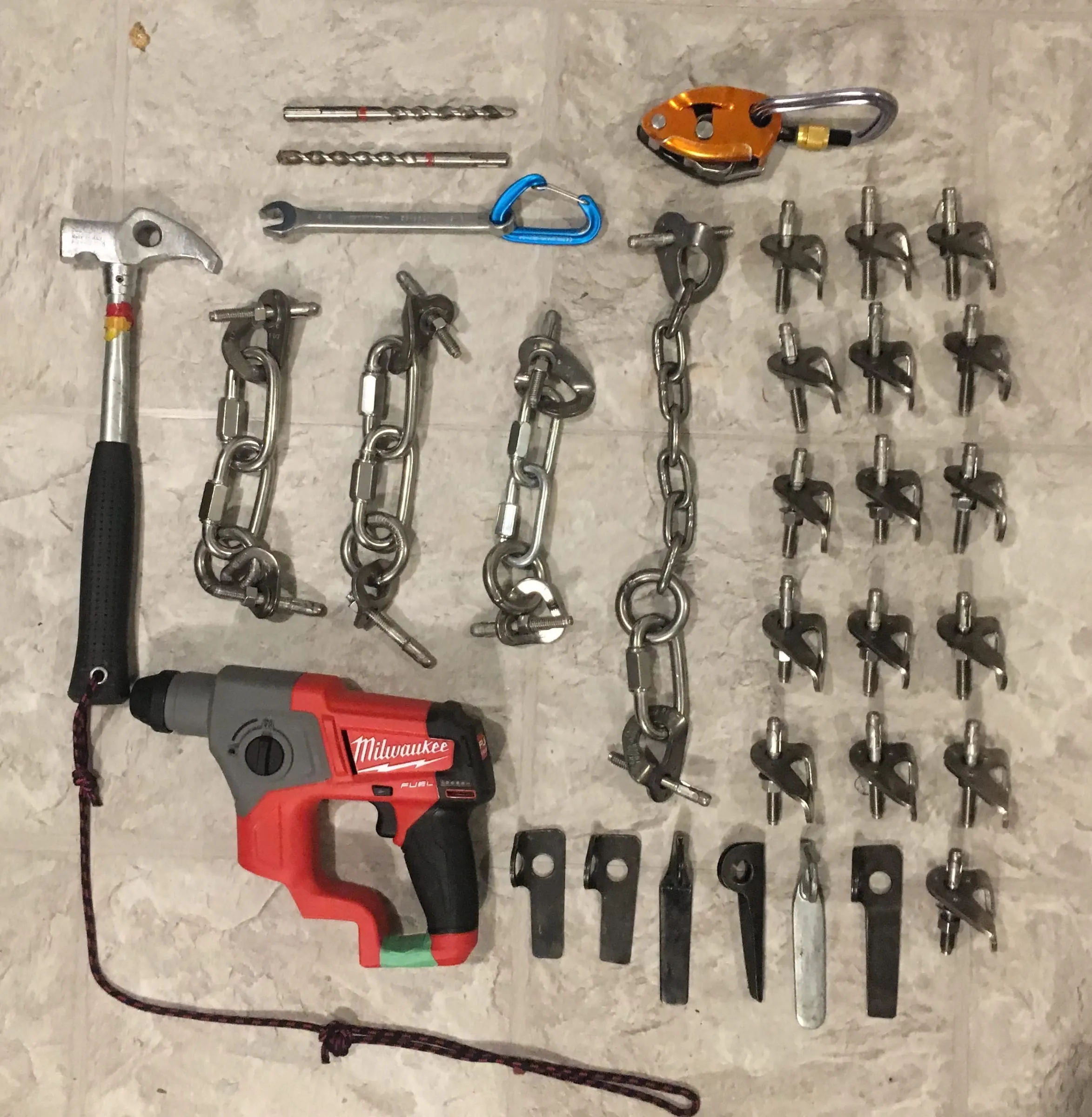 A retrofitting kit laid out on the floor: a rotary hammer, hammer, drill bits, wrench, belay device, stainless chain anchors, new bolts and hangers, and old hangers that have been removed