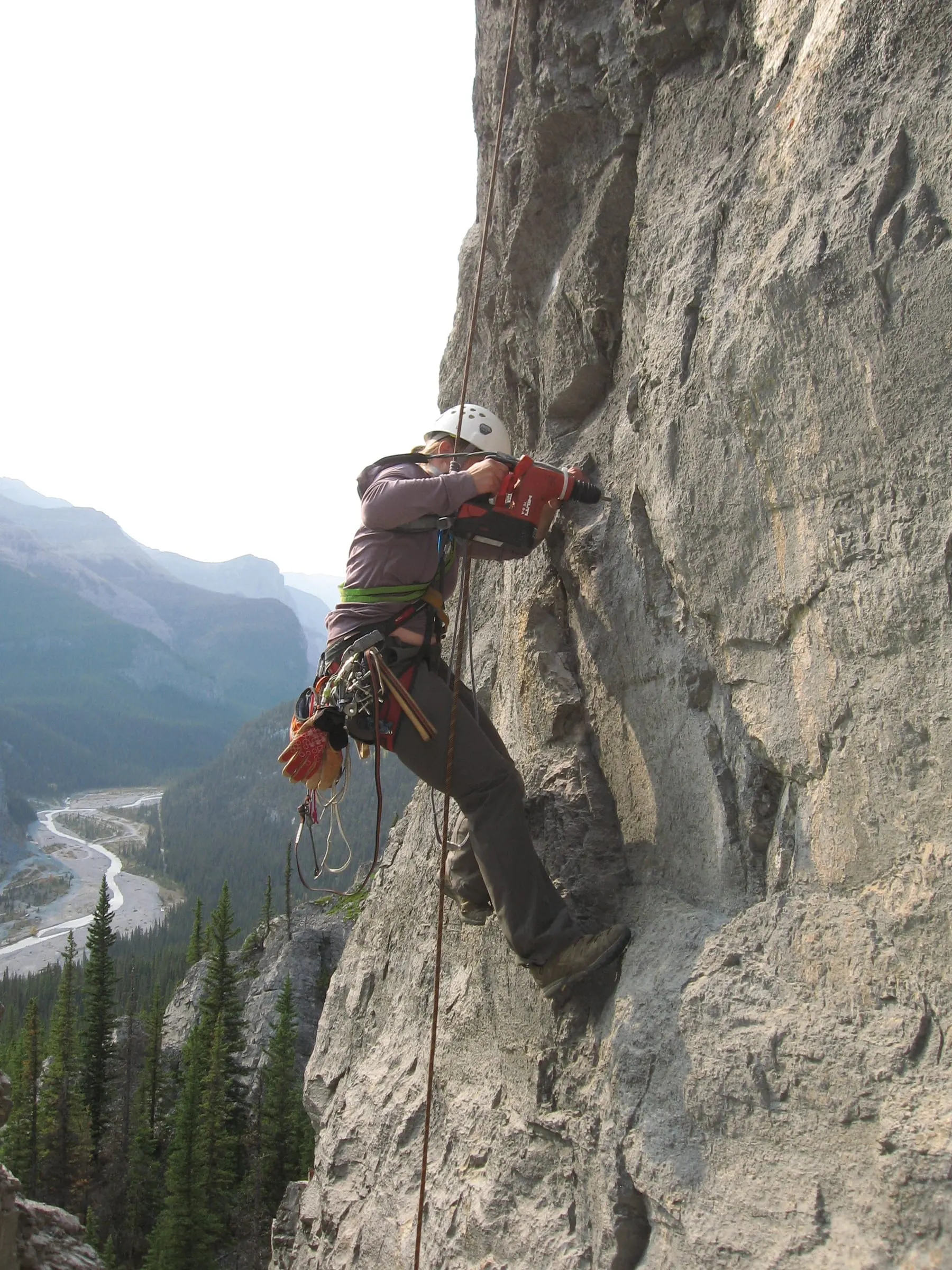 A route developer on rappel drills into a limestone wall high above a braided river valley